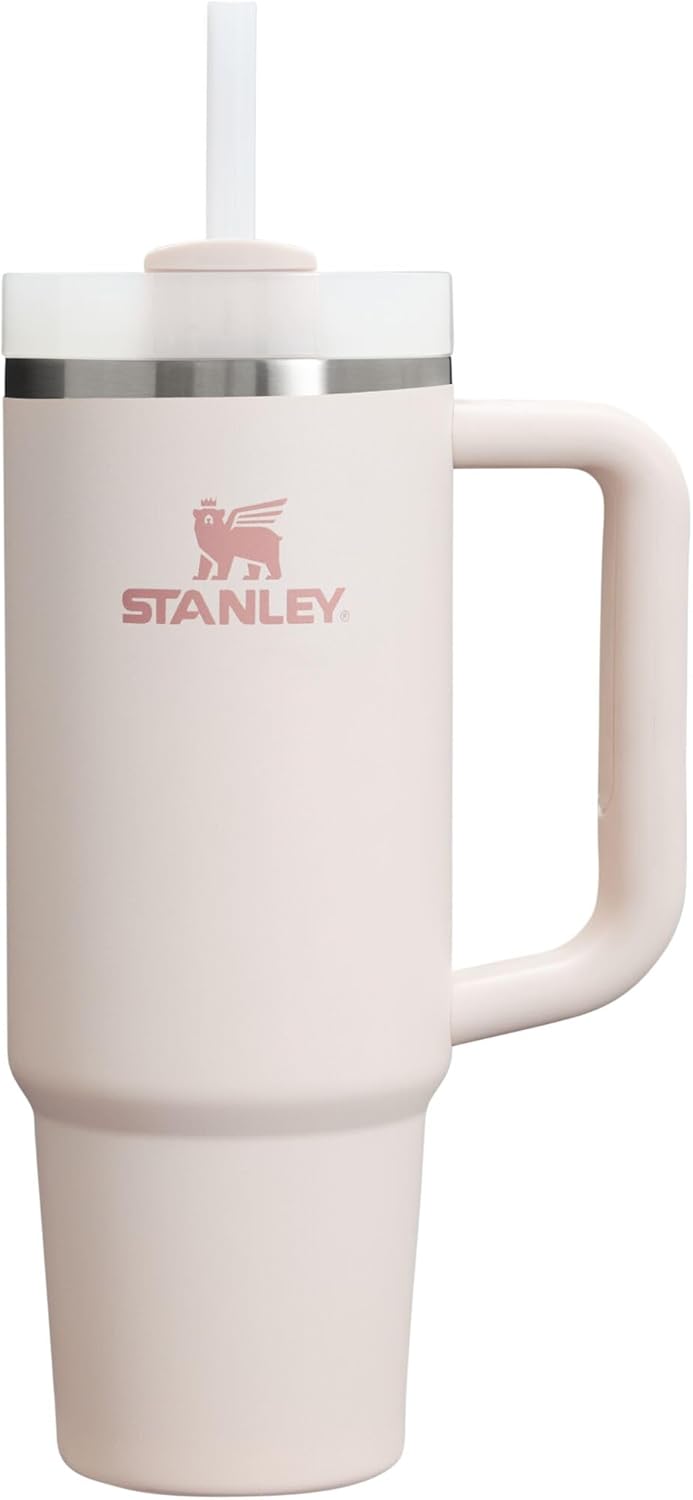 A Stanley insulated travel tumbler with a handle, a clear lid, and a straw. The tumbler is a very pale pink color with the Stanley brand logo printed in a matching dusty pink on the front, set against a plain white background.