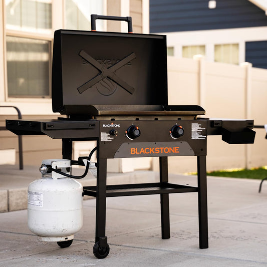 Black outdoor propane grill with open lid, side tables, two control knobs, and a white gas cylinder on a patio in front of a house.